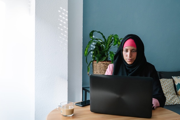 Giovane donna musulmana che indossa un hijab utilizzando la tecnologia al chiuso. Vista panoramica orizzontale della donna araba che lavora con il computer portatile a casa. Tecnologia e stile di vita delle donne musulmane.