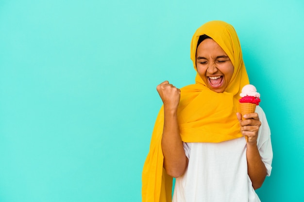 Giovane donna musulmana che mangia un gelato isolato sulla parete blu che alza il pugno dopo una vittoria, concetto del vincitore.