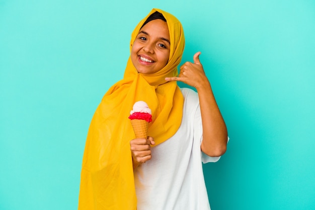 Giovane donna musulmana che mangia un gelato isolato sulla parete blu che mostra un gesto di chiamata di telefono cellulare con le dita.