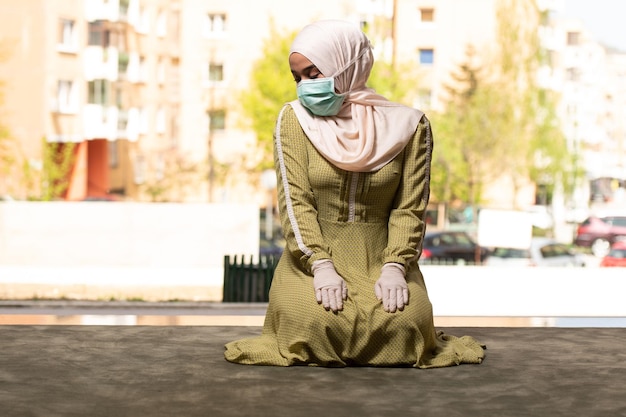 Giovane donna musulmana che prega all'aperto indossando maschera protettiva e guanti