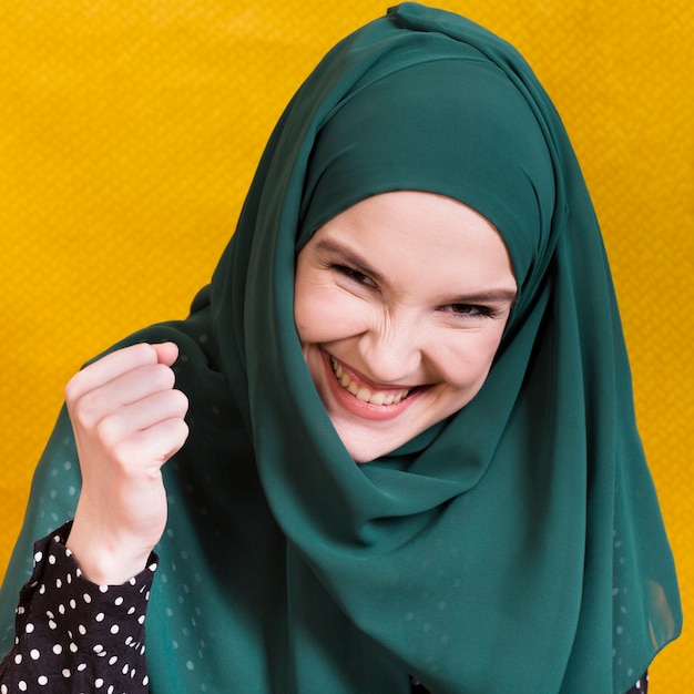 Giovane donna musulmana felice emozionante che esamina macchina fotografica davanti a fondo giallo