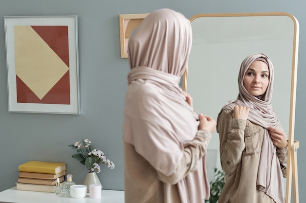 Giovane donna musulmana in hijab guardando in un grande specchio mentre si veste per il lavoro al mattino