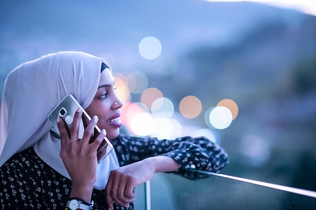 Giovane donna musulmana moderna che indossa il velo della sciarpa sulla strada urbana della città di notte mandando SMS sullo smartphone con la luce della città bokeh sullo sfondo