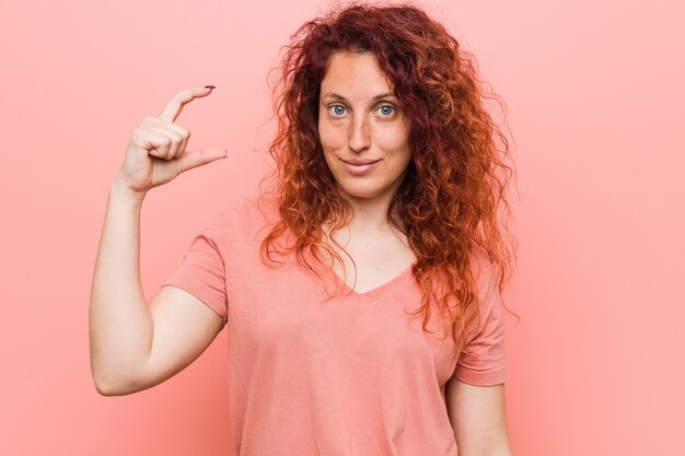 Giovane donna naturale e autentica rossa in possesso di qualcosa di piccolo con gli indici, sorridente e fiducioso.