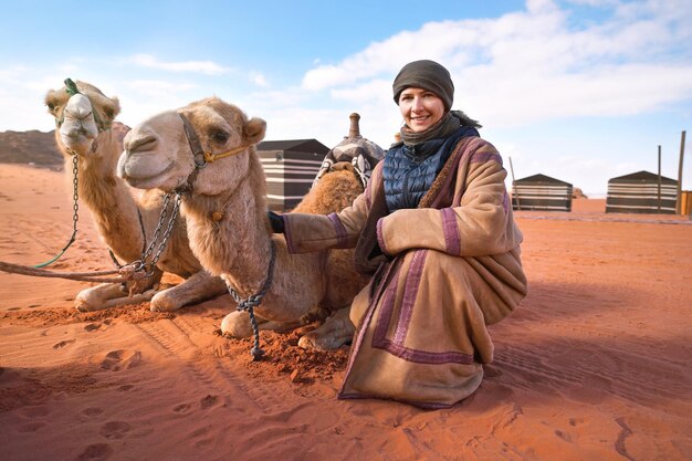 Giovane donna nel tradizionale cappotto beduino - bisht - e velo accovacciato accanto a due cammelli sdraiati sul deserto rosso terra, sorridente, tende da campo in background