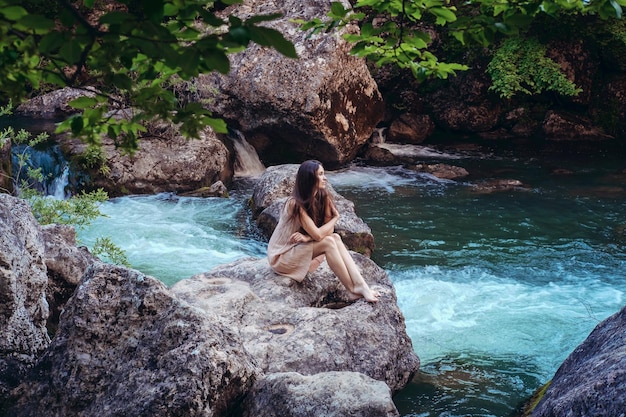 Giovane donna nel vestito è seduta sulla pietra nel mezzo di un torrente Tonalità alla moda