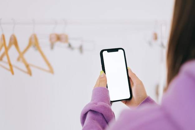 Giovane donna nell'utilizzo di smartphone con mock-up di schermo bianco nella stanza guardaroba bianco con cremagliera del pavimento.