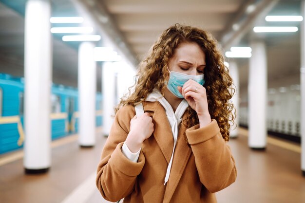 Giovane donna nella mascherina medica sterile protettiva che tossisce alla stazione della metropolitana.