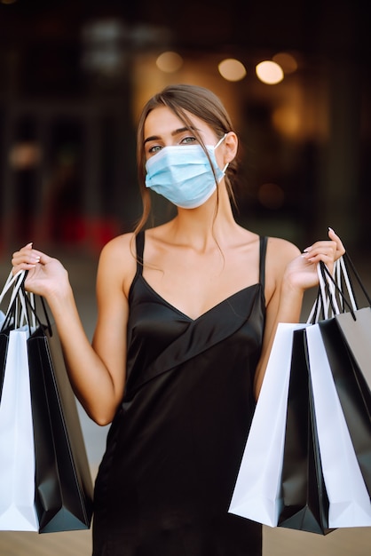 Giovane donna nella mascherina medica sterile protettiva sul suo fronte con i sacchetti della spesa che godono nello shopping.