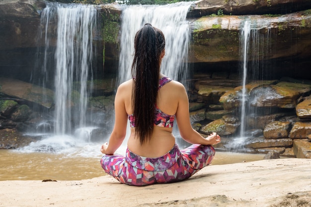 Giovane donna nella posa di yoga che si siede vicino alla cascata, retrovisione