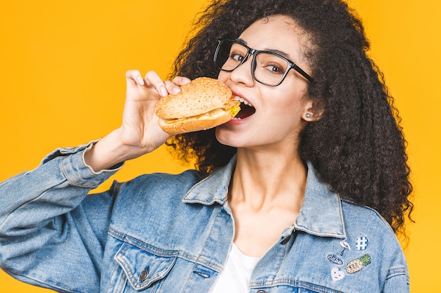 Giovane donna nera afroamericana che mangia hamburger isolato su fondo giallo.