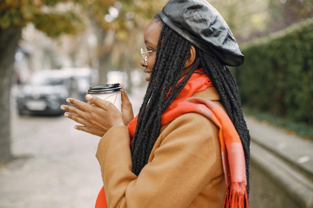 Giovane donna nera con lunghe acconciature locs in piedi fuori con una tazza di caffè da asporto. Donna che indossa cappotto marrone, sciarpa arancione e cappello nero