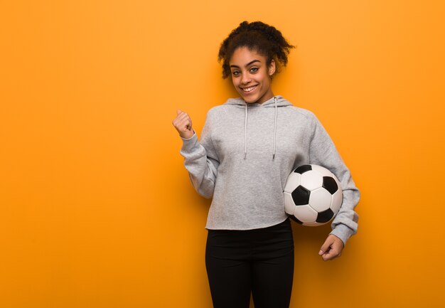 Giovane donna nera di forma fisica che sorride e che indica al lato. Tenendo un pallone da calcio.