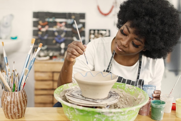 Giovane donna nera pittura vaso di terracotta presso lo studio d'arte Artigiana pittura ciotola di ceramica sul banco di lavoro Ragazza che indossa un grembiule blu