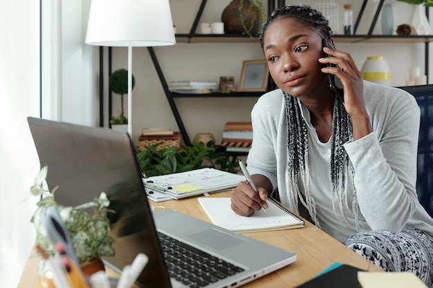 Giovane donna nera seria in abbigliamento da casa che parla al telefono con un collega, legge un documento sullo schermo del laptop e prende appunti