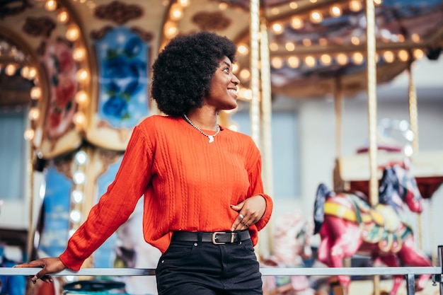 Giovane donna nera sognante sorridente con acconciatura afro e maglione rosso in piedi accanto a una giostra sulla strada