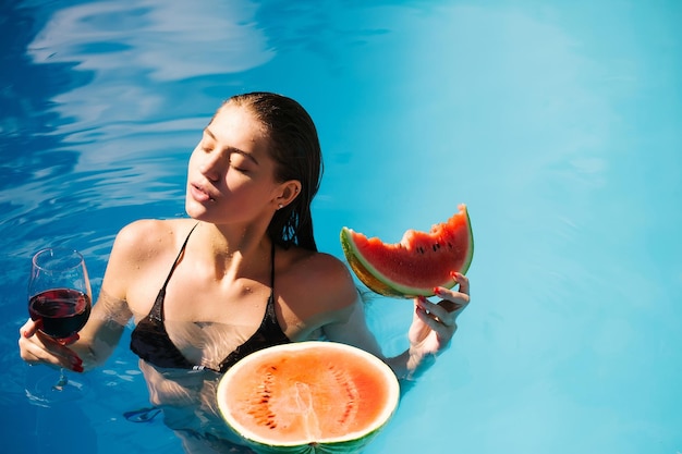 Giovane donna o ragazza sexy con bel viso e capelli bagnati, nuoto in piscina con acqua blu, mangiare anguria rossa e bere vino da vetro soleggiata giornata estiva all'aperto