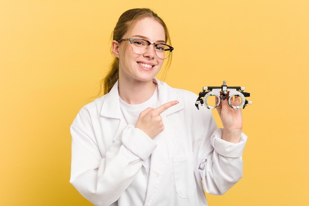 Giovane donna oculista caucasica che tiene un bicchiere di optometria isolato su sfondo giallo