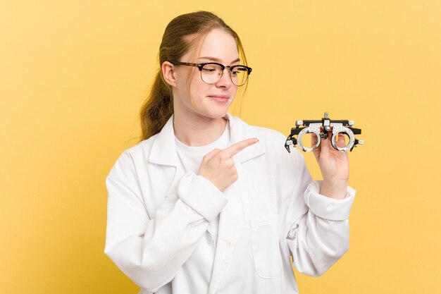 Giovane donna oculista caucasica che tiene un bicchiere di optometria isolato su sfondo giallo