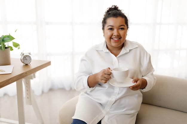 Giovane donna paffuta positiva che si siede alla scrivania che beve caffè