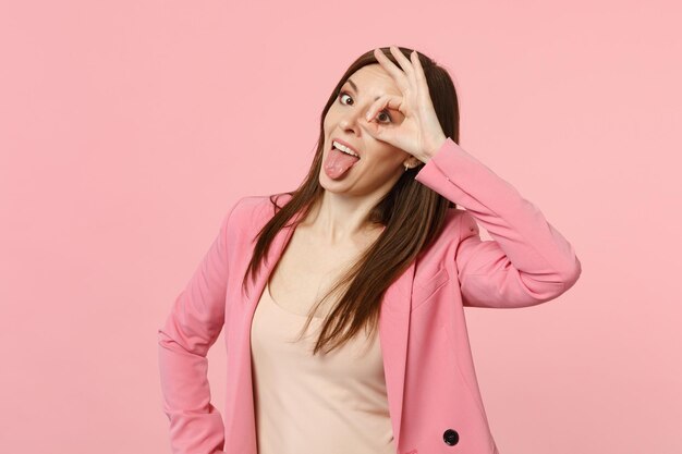 Giovane donna pazza in giacca che mostra la lingua, tenendo la mano vicino all'occhio, imitando occhiali o binocoli isolati su sfondo rosa pastello. Persone sincere emozioni, concetto di stile di vita. Mock up spazio di copia.