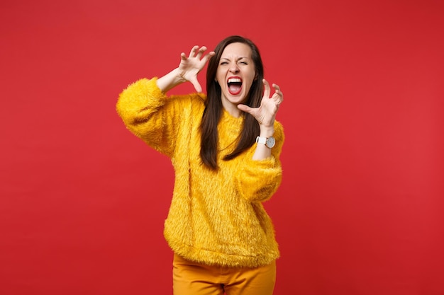 Giovane donna pazza in maglione di pelliccia gialla che grida, ringhiando come un animale, facendo gesto di artigli di gatto isolato su sfondo rosso in studio. Persone sincere emozioni, concetto di stile di vita. Mock up copia spazio.