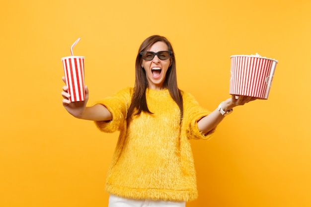 Giovane donna pazza in occhiali 3d imax che urla, guarda film, tiene in mano un secchio di popcorn, una tazza di plastica di cola o soda isolata su sfondo giallo. Persone sincere emozioni nel cinema, nello stile di vita.