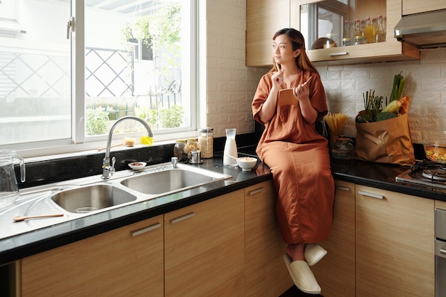 Giovane donna pensierosa che fa colazione, riempie il diario della gratitudine e guarda attraverso la finestra della cucina