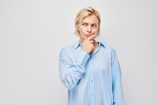 Giovane donna pensierosa con la mano sul mento che indossa una camicia blu isolata su uno sfondo bianco