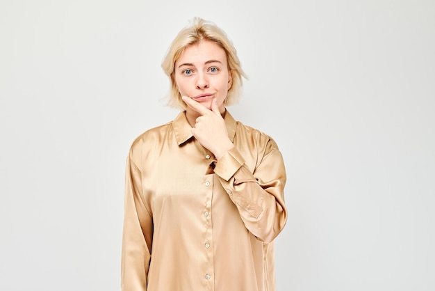 Giovane donna pensierosa con una camicia beige con la mano sul mento che sembra pensierosa su uno sfondo bianco