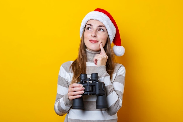 Giovane donna pensierosa in cappello di Babbo Natale con il binocolo su sfondo giallo