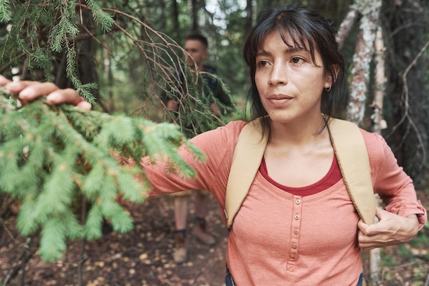 Giovane donna pensosa di razza mista con la borsa che tocca il ramo di un albero di abete mentre determina la direzione nella foresta