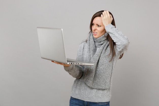 Giovane donna perplessa in maglione grigio, sciarpa che mette mano sulla testa lavorando su computer pc portatile isolato su sfondo grigio muro. Stile di vita sano, consulenza sul trattamento online, concetto di stagione fredda.