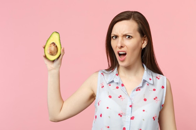 Giovane donna perplessa interessata in abiti estivi che tiene metà del frutto di avocado verde maturo fresco isolato su sfondo rosa pastello. Lo stile di vita vivido delle persone si rilassa il concetto di vacanza. Mock up spazio di copia.