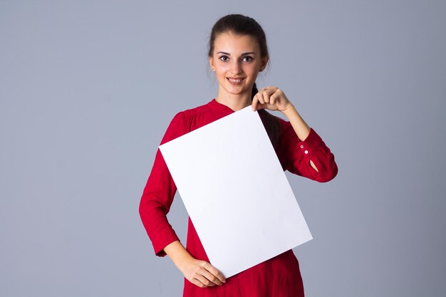 Giovane donna piacevole in camicetta con treccia che tiene foglio di carta bianco su sfondo grigio in studio