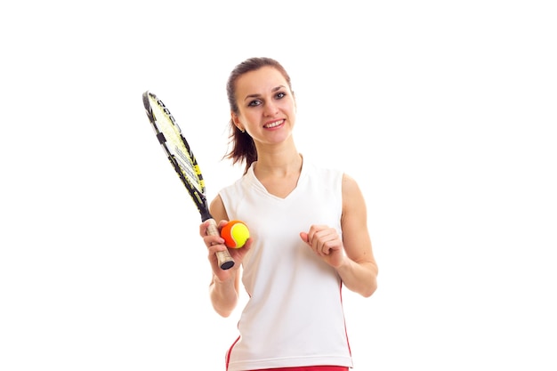 Giovane donna piacevole in camicia sportiva bianca con coda di cavallo scura che tiene racchetta da tennis e palla arancione su sfondo bianco in studio