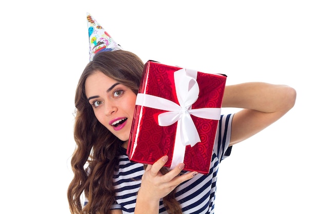 Giovane donna piacevole in maglietta spogliata e berretto da celebrazione che mostra un regalo rosso in studio