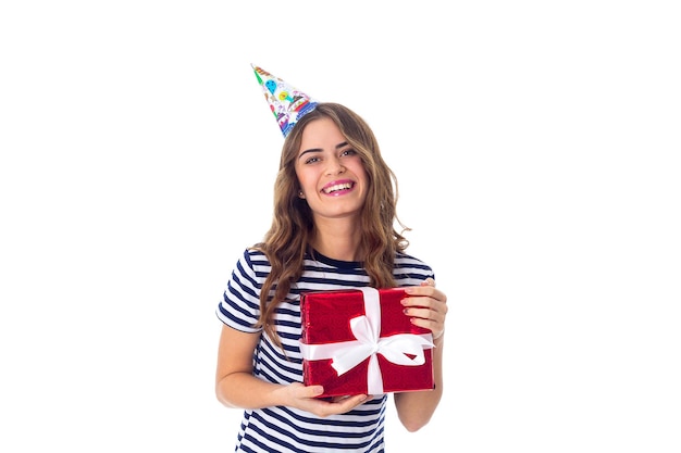 Giovane donna piacevole in maglietta spogliata e berretto da festa con capelli lunghi che tengono un regalo rosso su sfondo bianco in studio