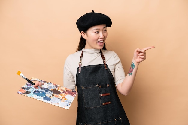 Giovane donna pittore cinese isolata su sfondo beige che punta il dito di lato e presenta un prodotto