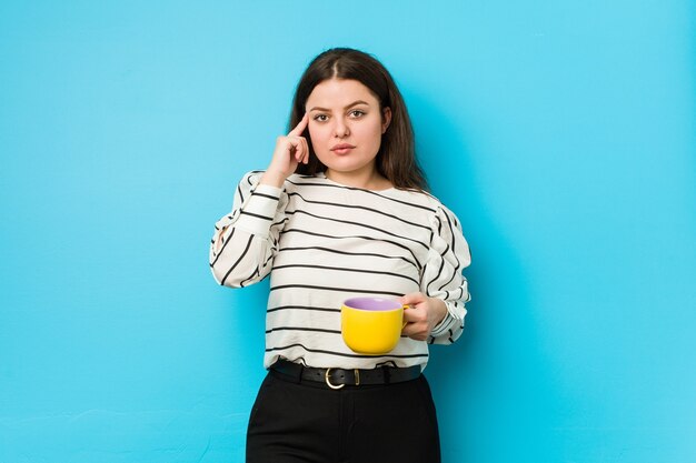 Giovane donna plus size che tiene una tazza di tè che punta la tempia con il dito, pensando, concentrata su un compito.