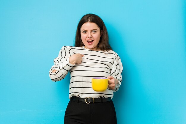 Giovane donna plus size in possesso di una tazza di tè sorpreso indicando se stesso, sorridendo ampiamente.