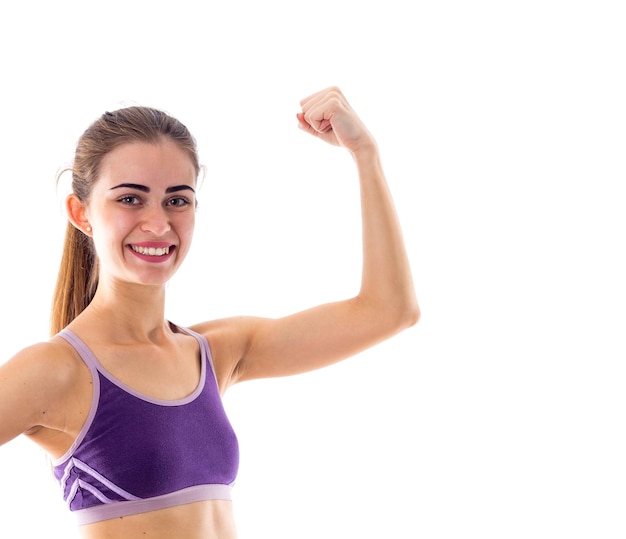 Giovane donna positiva che indossa un top sportivo viola con coda di cavallo che mostra i suoi bicipiti su sfondo bianco in studio