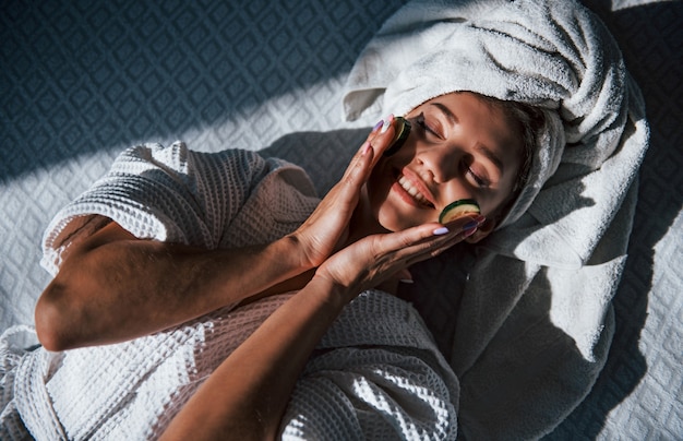 Giovane donna positiva con asciugamano sulla testa sdraiata sul letto con cetriolo.