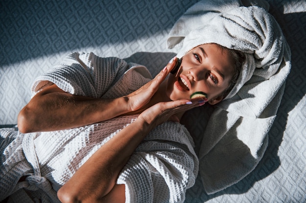 Giovane donna positiva con asciugamano sulla testa sdraiata sul letto con cetriolo.