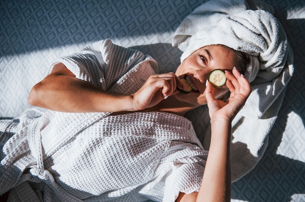 Giovane donna positiva con asciugamano sulla testa sdraiata sul letto con cetriolo.