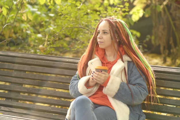 Giovane donna positiva con i dreadlocks allo zenzero che bevono caffè distogliendo lo sguardo seduto su una panchina nel parco in autunno dorato