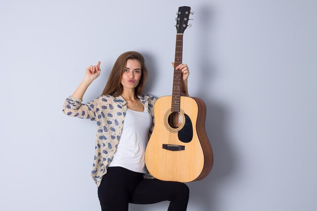 Giovane donna positiva in camicetta beige e pantaloni neri che tiene una chitarra su sfondo grigio in studio