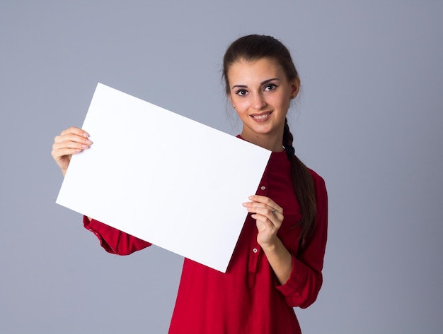 Giovane donna positiva in camicetta rossa con treccia che mostra un foglio di carta bianco su sfondo grigio in studio