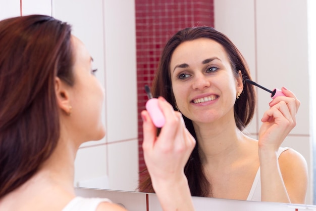 Giovane donna positiva in camicia con mascara rosa davanti allo specchio nel suo bagno bordeaux