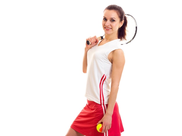 Giovane donna positiva in camicia sportiva bianca e gonna rossa che tiene una racchetta da tennis e una palla gialla